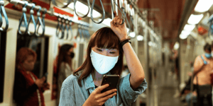 A woman on a train wearing a mask and looking at her phone she's holding with her hand, and the other holding on to the hand rail above her head.