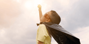 A little boy wearing a cape and sunglasses with his fist up in the air like a superhero.